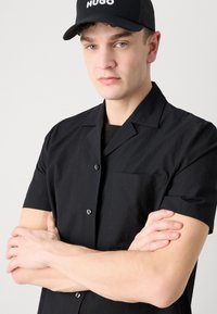 Trumpas juodas marškinėlių švarkas su tekstūruota medžiaga ir krūtinės kišene, derinamas su juoda kepure, ant kurios yra balti įrašai.