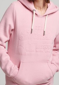 Sudadera rosa hecha de tela suave con un diseño de logo en relieve. Tiene una capucha con cordón y un bolsillo canguro para mayor funcionalidad.
