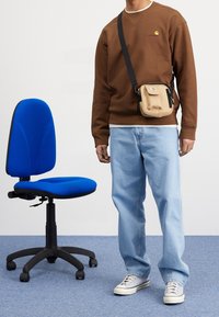 Brauner Hoodie mit Logo, hellblaue Jeans, beige Umhängetasche, graue Sneakers, neben einem blauen Bürostuhl auf einem strukturierten Boden.