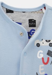 Hellblauer Baumwoll-Strampler mit Druck eines Vintage-Autos und einem farbenfrohen "GUESS" Schriftzug auf grauem Muster im Innenfutter.