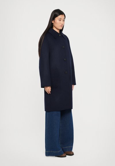 Marineblauwe wollen mantel met een grote kraag, voor knopen en zijzakken, gedragen met wijde blauwe denim broek en bruine schoenen.