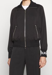 Giubbotto bomber nero con chiusura a zip anteriore, polsini e vita a coste. Presenta due tasche laterali con zip e testo bianco a contrasto sul polsino.