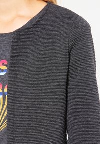 Grått, strukturerat tyg av en cardigan med ett subtilt ribbat mönster, med rund halsringning och öppen framsidesdesign. Någon grafisk detalj nedanför.