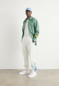 Grøn og blå ternet skjorte med knapper, hvid t-shirt, hvide sweatpants med grønne og blå striber, og hvide sneakers. Lyseblå kasket.