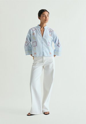Mujer con chaqueta bordada azul claro, pantalones blancos de pierna ancha y sandalias negras, de pie con las manos en los bolsillos frente a un fondo liso.