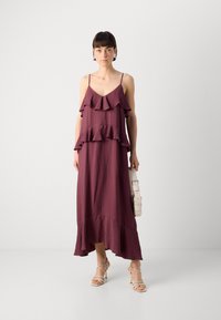 Bordeauxfarbenes ärmelloses Kleid mit gestuften Rüschen, einem V-Ausschnitt und einem asymmetrischen Saum aus strukturiertem Stoff; getragen mit Riemchenheels.
