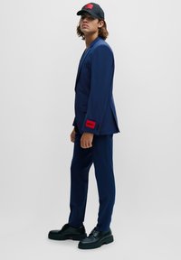 Marineblauer Anzug mit schlankem Design, versehen mit einem roten HUGO-Label am Ärmel. Eine schwarze Mütze und klobige schwarze Schuhe runden das Outfit ab.