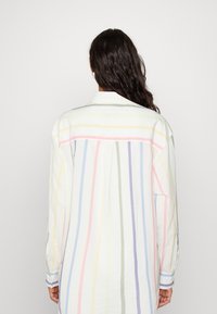 Femme aux longs cheveux noirs portant une chemise blanche ample avec des rayures verticales pastel, de dos contre un fond clair et uni.