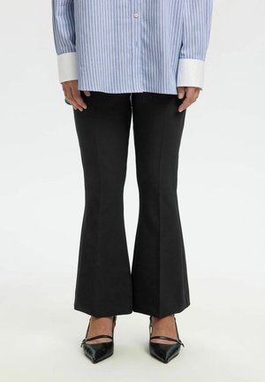 Személy fekete bővülő nadrágot, fekete hegyes orrú magassarkút és kék-fehér csíkos inget fehér mandzsettával visel, egyszerű háttér előtt áll.