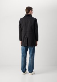 Cappotto lungo nero con colletto, caratterizzato da una texture liscia e un taglio dritto, abbinato a jeans azzurri chiari e sneakers bianche.