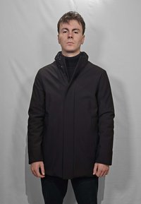 Giacca nera impermeabile con collo alto, chiusura frontale a scatto e tasche laterali. Indossata sopra un maglione scuro a maglia. Design semplice e pulito.