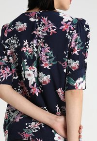 Blusa floreale con sfondo blu navy, maniche corte a palloncino e una varietà di fiori colorati. Tessuto texturizzato con una vestibilità rilassata.