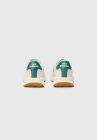 Krémové športové topánky s tyrkysovými akcentmi, zaoblený tvar, štruktúrovaná látka a gumové podrážky, s logom "New Balance" na päte.