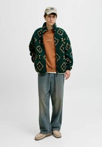 Giacca in pile verde a stampa con dettagli in beige, felpa marrone, pantaloni in denim larghi e scarpe slip-on beige. Cappellino con logo "W".