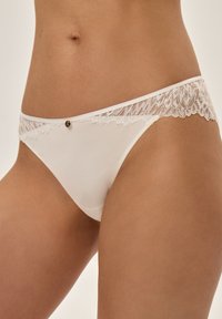 White bikini brief featuring a lace waistband and a small gold emblem at the front. Smooth fabric and delicate lace detailing.
