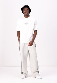 White cotton T-shirt with printed logo on chest, paired with light grey sweatpants. Model wearing a black bucket hat and white sneakers.