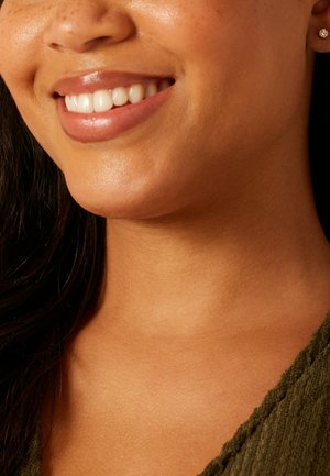 Close-up van het onderste deel van het gezicht en de nek van een glimlachende vrouw met een gladde huid, die een olijfgroen textuurshirt en een kleine oorbel draagt.