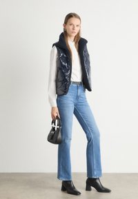 Gilet imbottito blu navy con colletto alto, indossato sopra una camicia bianca. Jeans azzurri e stivali a tacco nero. Piccola borsa nera tenuta in mano.