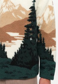 Fleece fabric with textured mountain and pine tree landscape pattern in beige, dark green, and black tones on clothing sleeve and torso.