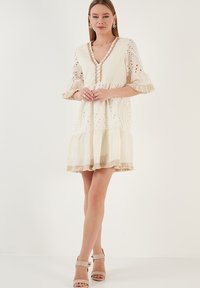 Robe blanche brodée avec des manches courtes à volants, décolleté en V profond, ourlet superposé et détails en dentelle. Associée à des sandales à talons beiges.