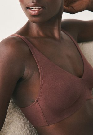 Femme noire portant un soutien-gorge sans fil mauve, penchée en arrière avec un bras levé derrière la tête, montrant une peau lisse et une posture détendue.