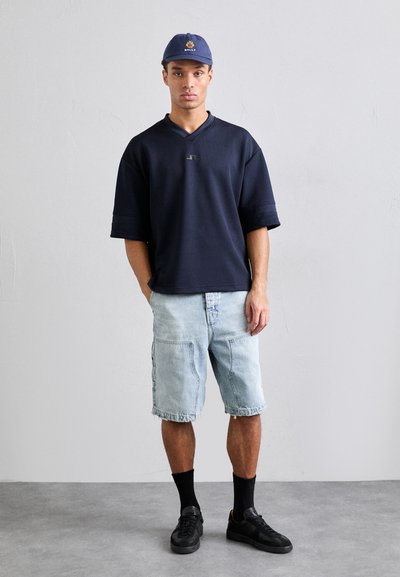 Marine oversized t-shirt med korte ærmer, lyse denim shorts med lommer, sorte sneakers og en marineblå kasket med et logo.