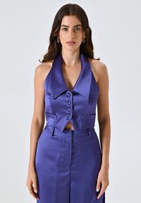 Gilet corto in satin viola con un ampio colletto, chiusura frontale con bottoni e tasche laterali, abbinato a pantaloni coordinati. Texture liscia.