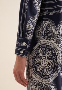 Camicia blu navy con motivo paisley bianco, dotata di polsini con bottone e tasca laterale. Tessuto liscio con dettagli intricati.