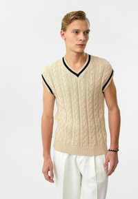 Beige kortemouwen gebreide pullover met kabelpatroon en V-hals. Heeft een zwarte geribbelde rand langs de halslijn en mouwen. Soepele textuur.