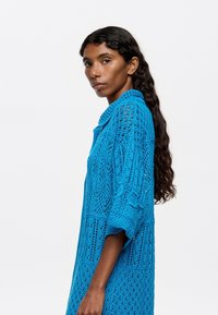 Abito in maglia di un blu vivace, caratterizzato da un design traforato, maniche oversize e colletto. La texture include motivi intricati in tutto l'abito.