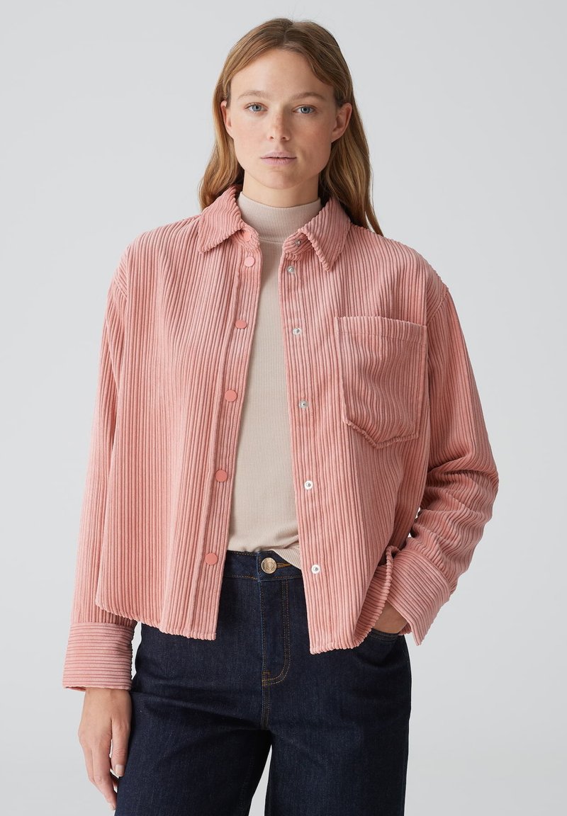 Pink corduroy shirt jacket with a button-up front, a single chest pocket, and a relaxed fit, paired with dark denim pants.