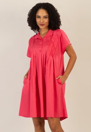 Woman with curly hair wearing a bright pink short-sleeve dress with pleats and pockets, standing against a plain beige background.