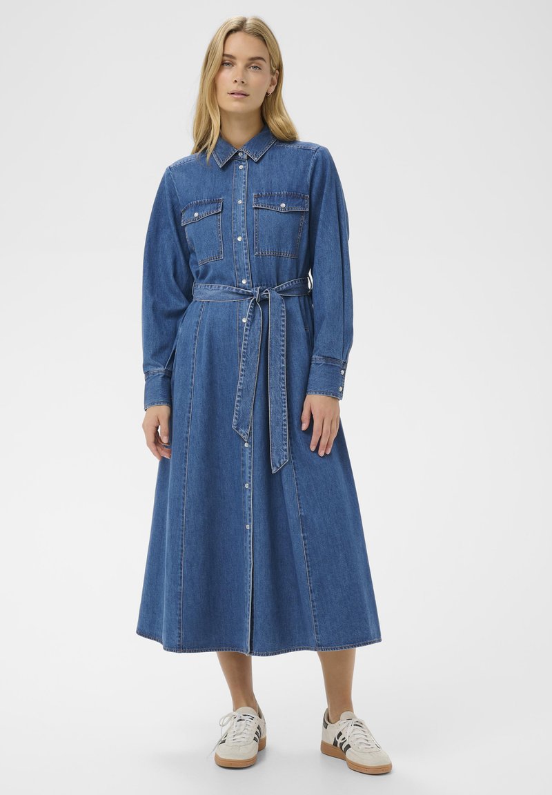 Jeanshemdjurk met lange mouwen, kraag, twee borstzakken, een ceintuur in de taille en een rok tot op de grond in blauw. Gestyled met witte sneakers.