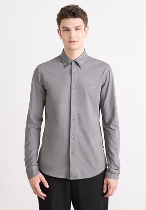 Camisa de botón de manga larga gris, con un cuello clásico, textura suave y un sutil logo en el pecho, acompañada de botones a juego.