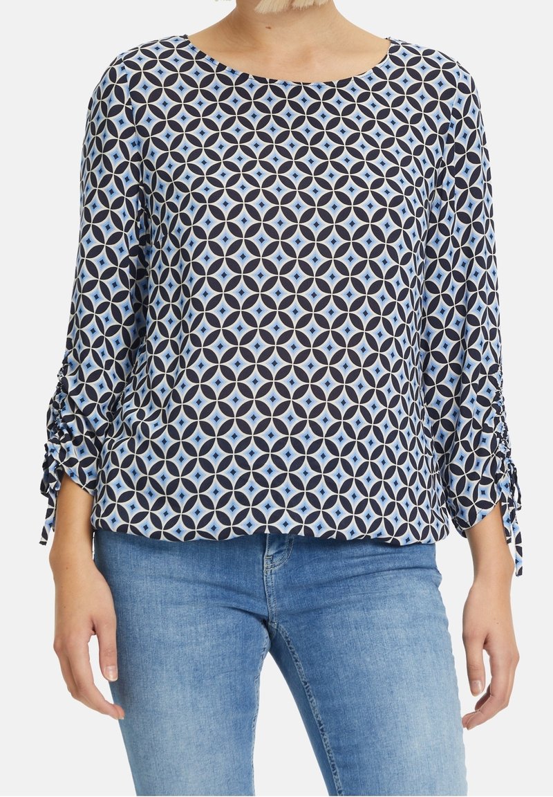 Blouse bleu marine avec un motif géométrique en bleu clair et crème. Dispose d'un col en rond et de manches légèrement froncées.