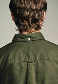 Chemise vert olive avec une texture douce, dotée d'une fermeture à boutons au col et d'un pli à l'arrière pour plus de détail et d'ajustement.