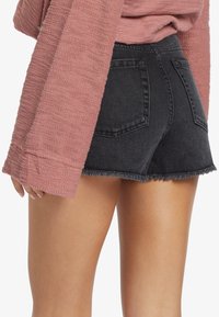 Pantalones cortos de mezclilla negra con un dobladillo deshilachado, combinados con una blusa de manga larga texturizada de color rosa. Los pantalones cortos tienen bolsillos curvados en la parte trasera.