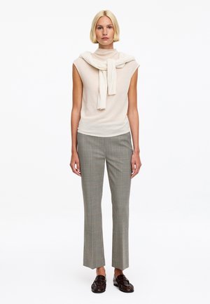 Femme blonde portant un haut beige sans manches avec un pull crème drapé sur les épaules, pantalon à carreaux et mocassins marron foncé, debout sur fond blanc.