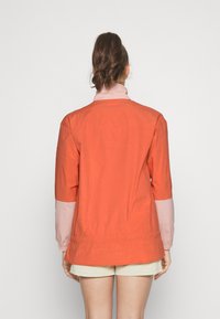 Kvinna med brunt hår i en hästsvans, iklädd en långärmad jacka i orange och ljusrosa samt beige shorts, står vänd bort.