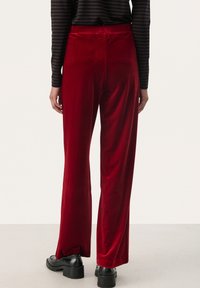 Velvet red trousers with wide legs, featuring a smooth texture and a high waistband. Paired with black platform shoes, viewed from the back.