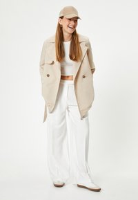 Trenca beige sobre un top corto blanco, pantalones anchos blancos y gorra beige. Las zapatillas presentan una combinación de colores crema y marrón.