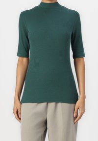 Femme portant un haut côtelé vert foncé à manches courtes et col montant, avec un pantalon beige clair à jambes larges, sur un fond uni.