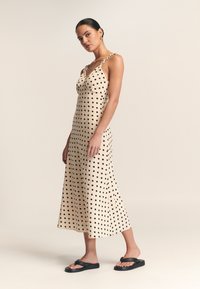 Midi-jurk van beige stof met zwarte stippen, voorzien van een diepe V-hals en gevlochten bandjes. Gepaarde met zwarte platte sandalen.