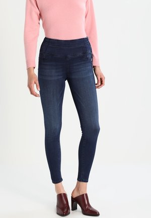 Vrouw draagt donkere skinny jeans, roze trui met lange mouwen en bordeauxrode slip-on schoenen met hak, staande tegen een witte achtergrond.