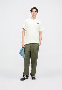 Bežs kokvilnas T-krekls ar īsām piedurknēm un logotipa printu, kombinēts ar olīvzaļām cargo biksēm un gaiši zilā cepurē, valkāts ar melnajām sporta apaviem.