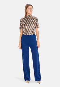 Multicolored patterned top with short sleeves and a high neckline, paired with high-waisted, wide-leg blue trousers. Smooth fabric texture.