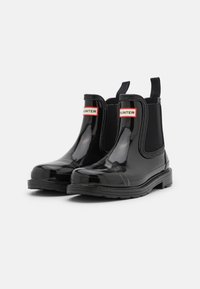 Zwarte rubberen Chelsea boots met glanzende afwerking, elastische zijpanelen en contrasterende rode en witte "HUNTER" branding op de enkel. Plat loopvlak.
