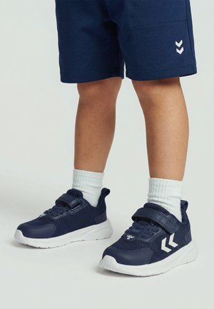 Kind draagt marineblauwe sportschoenen met witte zolen en witte sokken, gecombineerd met marineblauwe korte broek tegen een effen achtergrond.