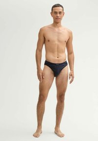 Schwarze Schwimmslips mit elastischem Bund, glatte Textur. Das Modell steht barfuß vor einem neutralen Hintergrund und präsentiert die Passform und das Design.