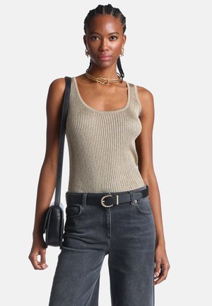Donna con capelli intrecciati che indossa un top a coste dorato, jeans neri con cintura, gioielli d'oro e porta una borsa a tracolla nera.